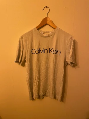 Vit Calvin Klein t-shirt - Säljer en klassisk vit t-shirt från Calvin Klein med stor svart logga över bröstet. T-shirten har rund halsringning och korta ärmar. Perfekt basic-plagg för dig som gillar stilrena och enkla looks.
