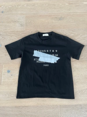 Svart Maison Margiela t-shirt -  svart t-shirt från Maison Margiela med ikoniskt tryck framtill i vitt och ljusgrått, samt texten 'Maison Margiela Paris'. Klassisk passform med rund hals och korta ärmar. Tillverkad i mjuk bomull för en skön känsla.