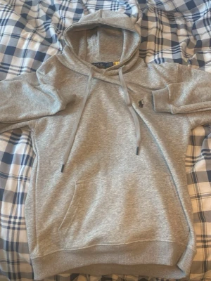 Grå hoodie från Polo Ralph Lauren - Snygg grå hoodie från Polo Ralph Lauren med klassisk huva, dragsnören och känguruficka. Liten broderad logga på bröstet. Tillverkad i mjukt bomullsmaterial som är perfekt för chill dagar. Enkel och stilren design som funkar till det mesta.är storlek S men passar om man har XS kom gärna med ett prisförslag 