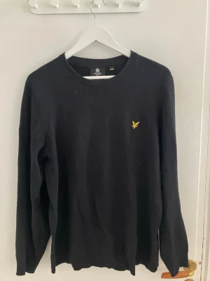 Svart stickad tröja från Lyle & Scott - Klassisk svart stickad tröja från Lyle & Scott med rund halsringning och den ikoniska gula örnloggan broderad på bröstet. Tröjan har långa ärmar och ribbade muddar vid ärmslut och nederkant. Perfekt för lager på lager under kyliga dagar.