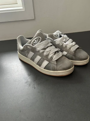 Adidas campus grå - Snygga grå sneakers från Adidas med klassiska vita ränder på sidorna och vit logga på plösen. Skorna har breda vita snören och en låg profil. Ovandelen är i mocka och textil, med en vit sula som har en brun kant. Perfekt för dig som gillar stilrena och sportiga skor.