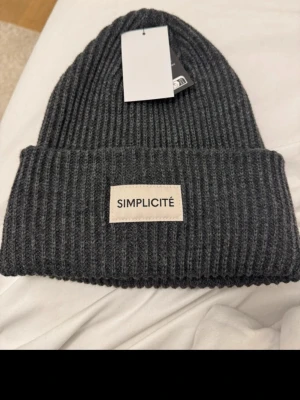 Mörkgrå ribbstickad mössa Simplicité - Snygg mörkgrå ribbstickad mössa från Simplicité med uppvikt kant och en beige patch med loggan framtill. Klassisk form och mjukt material som håller dig varm under kalla dagar. Perfekt accessoar för en stilren look.