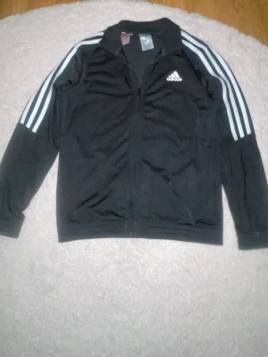 Svart Adidas kofta - Säljer en svart träningsjacka från Adidas med dragkedja framtill och klassiska vita ränder längs ärmarna. Jackan har hög krage och Adidas-logga på bröstet. Perfekt för sport eller chill. Tillverkad i ett mjukt och lätt material.