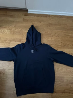 Mörkblå hoodie från Lacoste - Snygg mörkblå hoodie från Lacoste med klassisk huva och känguruficka framtill. Liten vit Lacoste-logga på bröstet och den ikoniska gröna krokodilen på ärmen. Tillverkad i mjukt bomullsmaterial som känns skönt mot huden. Perfekt för en avslappnad och stilren look.