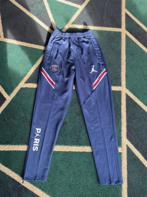 Blå PSG x Jordan träningsbyxor - Snygga blå träningsbyxor från Jordan x PSG med vita och röda detaljer vid låren. Byxorna har elastisk midja, logotyper på båda benen och texten 'PARIS' längs ena benet. Perfekta för träning eller chill.