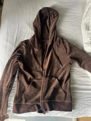 Brun Juicy Couture hoodie med fickor - Mysig brun hoodie med dragkedja framtill och stora kängurufickor. Tröjan har huva med dragsko och ribbade muddar vid ärmslut och nederkant. Perfekt för en avslappnad och chill stil. Materialet känns mjukt och bekvämt.