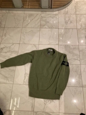 Grön stickad tröja från Stone Island - Snygg grön stickad tröja från Stone Island med klassisk patch på ärmen. Tröjan har rund halsringning, ribbade muddar och är tillverkad i mjukt material som passar perfekt till höst och vinter. Enkel men stilren design som funkar till mycket.