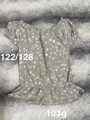 Beige blommig blus med volang - Söt beige blus med vitt blommönster och volangkant nedtill. Blusen har korta puffärmar med resår och en rundad halsringning med resår för en bekväm passform. Perfekt för en somrig look!🌸 Fint använt skick. Storlek 122/128