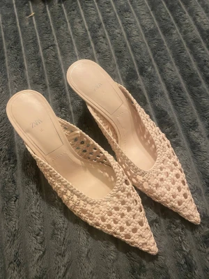 Beige skinnflätade klckar från Zara - Säljer ett par snygga beige klackar från Zara med spetsig tå och öppen häl. Skorna har ett flätat mönster i syntetmaterial som ger en luftig och trendig look. Perfekta för dig som gillar stilrena och moderna detaljer. Passar även 37. Helt nya
