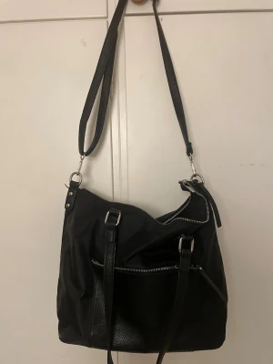 Svart axelväska med dragkedjor - Snygg svart axelväska med både lång och kort rem, flera dragkedjor och praktiska fack. Väskan har en stilren design i konstläder och tyg, perfekt för dig som vill ha plats för allt du behöver. Silverfärgade detaljer och justerbar axelrem.