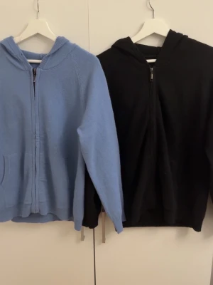 Kashmir hoodie  - Två hoodies i mjukt stickat material, en i ljusblått och en i svart. Båda har dragkedja framtill, huva och fickor på sidorna. Perfekta för en avslappnad och chill stil. Passar till jeans eller mjukisbyxor för en enkel vardagslook.