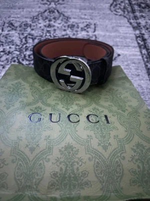 Gucci svart bälte med silver GG-spänne - Snyggt svart bälte från Gucci med det ikoniska GG-spännet i silver. Bältet har en klassisk design med diskret mönster på lädret och insida i brunt. Perfekt accessoar för att lyfta din outfit.