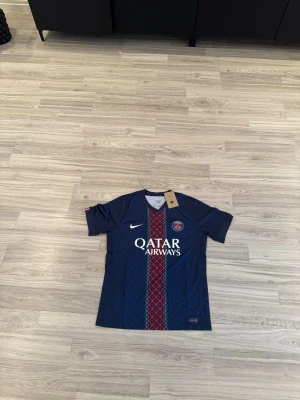 PSG matchtröja 2025/26  - Snygg blå Paris Saint-Germain matchtröja från Nike med röd och vit detalj i mitten och PSG-logga på bröstet. Tröjan har Qatar Airways-tryck framtill och är tillverkad i ett lätt, ventilerande material perfekt för fotboll eller träning.