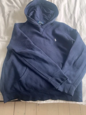 Mörkblå hoodie från Polo Ralph Lauren - Klassisk mörkblå hoodie från Polo Ralph Lauren med huva och känguruficka. Diskret broderad logga på bröstet och mjukt material som känns skönt mot huden. Perfekt för en avslappnad och stilren look.