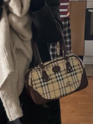 Burberry hand väska - Snygg handväska med klassiskt beige, rött och svart rutmönster och bruna läderdetaljer. Väskan har två handtag och silverfärgade nitar. Perfekt för dig som gillar en tidlös och trendig look.