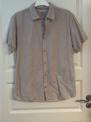 Beige kortärmad skjorta Essentials - Luftig beige skjorta från Essentials med korta ärmar och klassisk krage. Skjortan har knappar framtill och är tillverkad i mjuk bomull, perfekt för varma dagar. Enkel och stilren design som passar till det mesta.