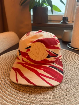 Moncler keps med rödvit mönster - Säljer en snygg keps från Moncler med ett unikt rödvitt mönster och svarta detaljer. Kepsen har en stor rund Moncler-logga framtill och är tillverkad i bomull. Perfekt för dig som vill sticka ut med en trendig accessoar.