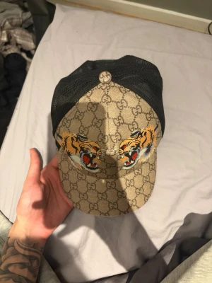 Gucci keps med tigertryck och GG-mönster - Säljer en snygg Gucci keps med klassiskt beige GG-mönster och två stora, färgstarka tigerhuvuden framtill. Kepsen har svart mesh baktill för extra ventilation och justerbar passform. Perfekt för dig som vill sticka ut med en exklusiv accessoar.