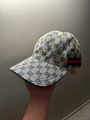 Gucci beige keps med GG-mönster - Snygg beige keps från Gucci med klassiskt blått GG-monogram över hela kepsen. På sidan finns en ikonisk röd och grön rand. Kepsen har böjd skärm och är tillverkad i canvasmaterial som ger en exklusiv känsla. Perfekt för dig som vill sticka ut med en lyxig accessoar. Finns i Malmö skriv vid intresse!