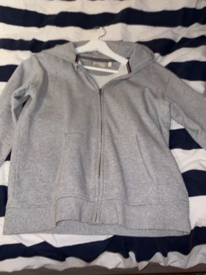 Grå zip hoodie från GANT - Snygg grå hoodie från GANT med dragkedja framtill och två fickor. Klassisk design med ribbade muddar och huva. Insidan är mjuk och bekväm, perfekt för chill dagar. Detalj med randig kant på insidan av kragen ger en sportig touch.