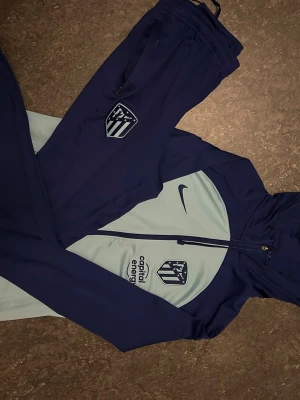 Nike Atlético Madrid träningsset blå - Snyggt träningsset från Nike i mörkblått och ljusblått med Atlético Madrid-logga och Capital Energy-tryck. Setet består av ziptröja med huva och matchande byxor, båda med klubbmärke. Perfekt för fotbollsträning eller chill.