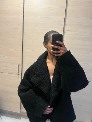 Svart oversized teddyjacka Gina tricot - Mysig svart teddyjacka med oversized passform och stora slag. Jackan har en fluffig, mjuk struktur och stängs med knappar framtill. Perfekt för att hålla sig varm och stilren under kyliga dagar. 