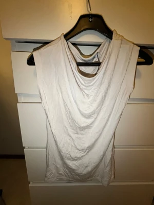 Vit ärmlös topp med draperad hals - Snygg vit topp med draperad halsringning och ärmlös design. Perfekt för dig som gillar en enkel men stilren look. Tillverkad i mjukt material som känns skönt mot huden. Passar till många olika outfits.