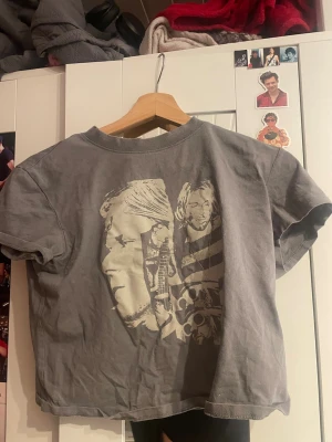 Grå t-shirt med kurt cobain tryck - Cool grå t-shirt med ett stort tryck på kurt cobain på framsidan. T-shirten har korta ärmar och en klassisk rund hals. Perfekt för dig som gillar streetstyle och unika motiv. T-shirten är lite som en magtröja och börjar ungefär vid naveln.