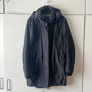 Svart vinterjacka  - Säljer en svart vinterjacka från Didriksson med huva och dragsko i midjan och längst ner. Jackan har långa ärmar, knäppning med både dragkedja och knappar samt två snedställda fickor framtill. Perfekt för kyligare dagar och enkel att styla till olika outfits. 