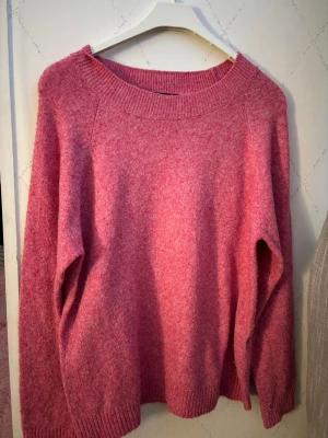 Rosa stickad tröja med bred halsringning - Mysig rosa stickad tröja med bred ribbad halsringning och långa ärmar. Tröjan har en avslappnad passform och ribbade muddar vid ärmslut och nederkant. Perfekt för att hålla värmen under kyliga dagar.