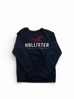 Mörkblå Hollister sweatshirt - Mörkblå sweatshirt från Hollister med stort tryck framtill i vitt och rött. Klassisk rund halsringning och långa ärmar. Perfekt för en avslappnad och stilren look. Tjockt och mjukt material som håller dig varm.