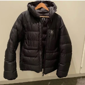 Svart dunjacka från Haglöfs - Snygg svart dunjacka från Haglöfs med huva och dragkedja framtill. Jackan har två sidofickor med dragkedja och diskret Haglöfs-logga på bröstet. Perfekt för kalla vinterdagar tack vare det tjocka dunfodret och den klassiska quiltade designen.
