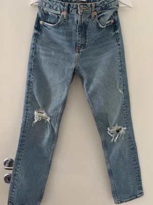  Blå jeans med raka ben och slitningar från Zara - Säljer ett par klassiska blå jeans från Zara med raka ben och slitna detaljer vid knäna. Jeansen har fem fickor, hög midja och är gjorda i kraftig denim. Perfekta för en avslappnad och trendig look.