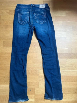 True Religion blå bootcut jeans - Säljer ett par klassiska blå jeans från True Religion med bootcut-modell. Jeansen har tydliga kontrastsömmar och ikoniska bakfickor med broderad logga. Tillverkade i denim med en snygg tvätt och normal midja. Perfekta för en avslappnad och trendig look. Storlek 29 