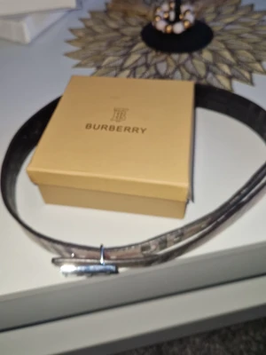 Rutig Burberry-bälte med TB-spänne - Snyggt bälte från Burberry med klassiskt rutigt mönster i beige, svart och rött. Bältet har ett stilrent metallspänne med Burberrys TB-logga framtill. Perfekt accessoar för att lyfta din outfit.