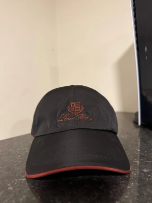 Svart keps från Loro Piana - Säljer en svart keps från Loro Piana med röd broderad logotyp och emblem framtill. Kepsen har en böjd skärm med röd kant. Materialet är slätt och känns exklusivt, perfekt för dig som vill ha en stilren accessoar.