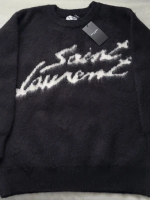 Svart Saint Laurent stickad tröja - Cool svart stickad tröja från Saint Laurent med vit logotyp i stor, handskriven stil på bröstet. Tröjan har rund halsringning och är gjord i ett fluffigt, mjukt material som känns lyxigt och trendigt. Perfekt för dig som vill ha en statement piece i garderoben.