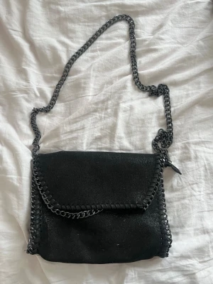 Svart axelväska med kedjerem från design by amour - Snygg svart axelväska med grov kedjerem och dekorativa kedjedetaljer längs kanterna. Väskan har en lockstängning och är tillverkad i ett material som liknar mocka. Perfekt för dig som gillar en edgy och trendig stil.