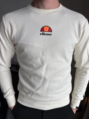 Vit sweatshirt från Ellesse - Säljer en stilren vit sweatshirt från Ellesse med klassisk logga i orange och rött på bröstet. Tröjan har rund halsringning, långa ärmar och ribbade muddar vid ärmslut och nederkant. Perfekt för en avslappnad och sportig look.