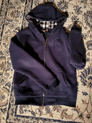 Mörkblå hoodie från Burberry - Snygg mörkblå hoodie från Burberry med dragkedja och klassiskt rutigt foder i huvan. Hoodien har fickor framtill och ribbade muddar vid ärmar och nederkant. Perfekt för dig som gillar stilrena detaljer och exklusiva märken.