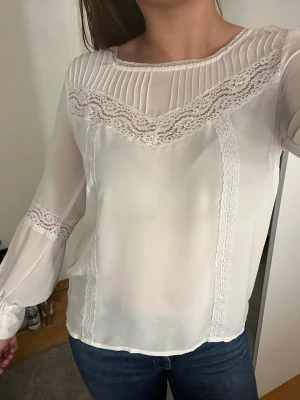 Vit blus med spetsdetaljer - Säljer en vit blus med långärmad design och fina spetsdetaljer längs bröst, ärmar och framstycke. Blusen har plisserad kant upptill och är gjord i ett tunt, lätt material som ger en elegant look. Perfekt att matcha med jeans eller kjol. 