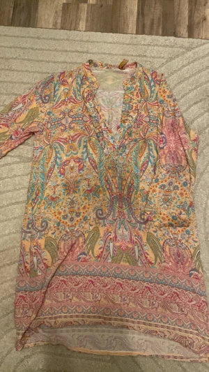 Mönstrad klänning i pastellfärger - Säljer en tunn och luftig blus med färgglatt paisleymönster i rosa, turkos, gult och orange. Blusen har v-ringning med små volangdetaljer och knappar framtill. Materialet känns lätt och flowy, perfekt för varma dagar. Ingen synlig logga eller märke. Söndring märkeslapp går enkelt att fixa.