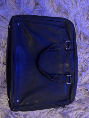 Svart portfölj från Le Tanneur - Snygg svart portfölj i äkta läder från Le Tanneur. Klassisk rektangulär form med dubbla handtag och silverfärgade metalldetaljer. Perfekt för laptop och dokument, stilren och tidlös design som passar till alla tillfällen. Nypris ish 4-5 tusen. Vill bara bli av med den så lägg ditt eget bud annars är de det pris som står😊