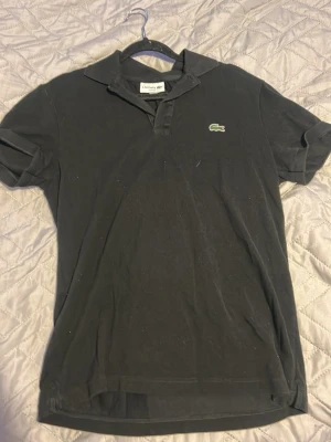 Svart pikétröja från Lacoste - Klassisk svart pikétröja från Lacoste i slim fit-modell. Tröjan har korta ärmar, krage och knappar framtill samt den ikoniska gröna krokodilloggan på bröstet. Perfekt för en clean och stilren look.
