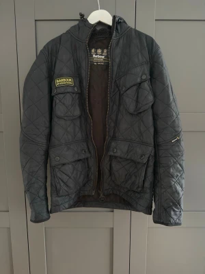 Svart quiltad jacka från Barbour - Snygg svart quiltad jacka från Barbour International med huva och flera praktiska fickor framtill. Jackan har dragkedja och patchdetalj på bröstet. Perfekt för dig som gillar klassisk men sportig stil.