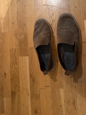 Bruna mockatofflor med gummisula - Mysiga tofflor i brun mocka med mjuk insida och slitstark gummisula. Perfekta att glida runt i hemma. Klassisk slip-in modell med diskret söm runt kanten. Passar dig som gillar enkel och stilren design.