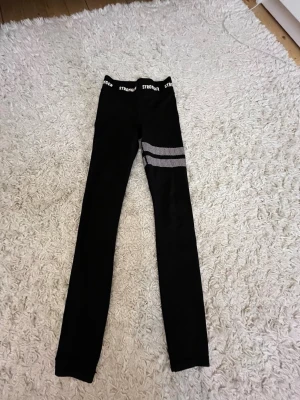 Svarta leggings från Stronger - Snygga svarta leggings från Stronger med vita detaljer i form av två randiga mönster på ena benet och logga i midjan. Tight passform och stretchigt material som passar perfekt till träning eller chill. Midjan är elastisk för extra komfort.