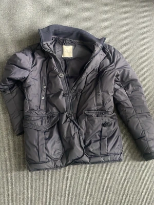 Dunjacka från Moncler - Snygg svart dunjacka från Moncler med hög krage, dragkedja och knappar framtill. Jackan har två stora fickor med lock och knappstängning samt quiltad design. Perfekt för kalla vinterdagar och riktigt stilren look. Storleken är size 2 vilket motsvarar S/M! Fler frågor eller funderingar skriv i dm!