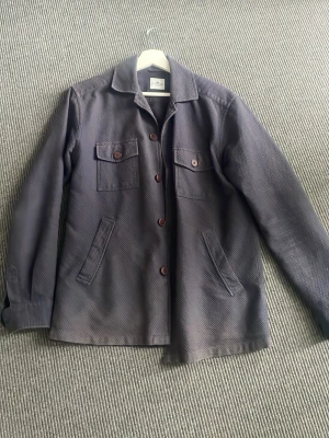 Mörkblå overshirt från John Henric - Stilren mörkblå overshirt från John Henric i strukturerat bomullstyg. Klassisk krage, fyra fickor framtill och bruna knappar. Perfekt lager-på-lager-plagg med avslappnad passform och snygga detaljer. Fler frågor eller funderingar hör av er i Dm!