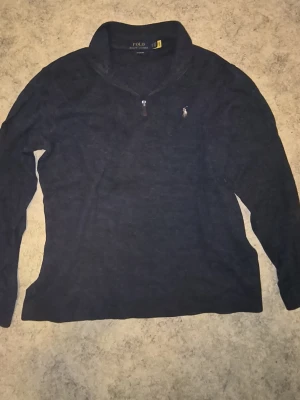 Svart stickad tröja Polo Ralph Lauren - Svart stickad tröja från Polo Ralph Lauren med halv dragkedja och klassisk broderad logga på bröstet. Tröjan har hög krage och långa ärmar, perfekt för lager på lager. Materialet känns mjukt och bekvämt.